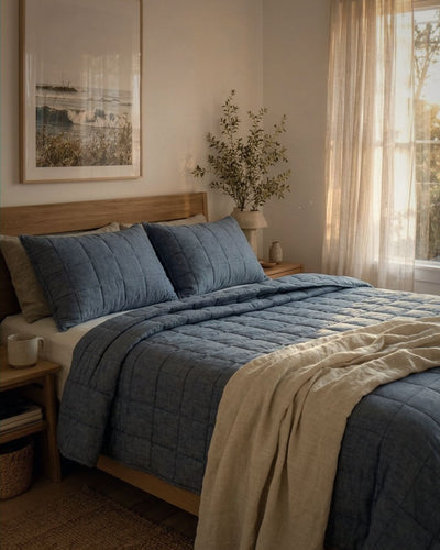 Bedroom with a bed featuring blue bedding and beige blanket, framed picture on the wall, and window with curtains.