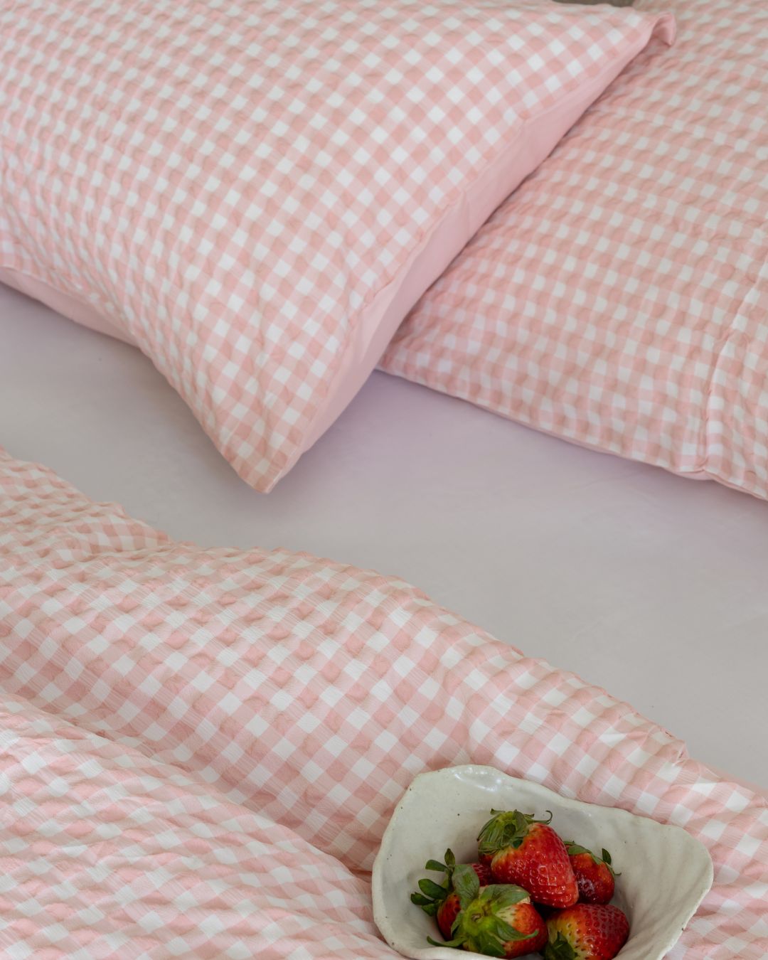 Pink checkered bedding with a bowl of strawberries on a light pink background