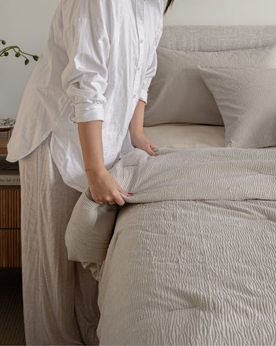 Person making a bed with beige bedding in a bedroom setting