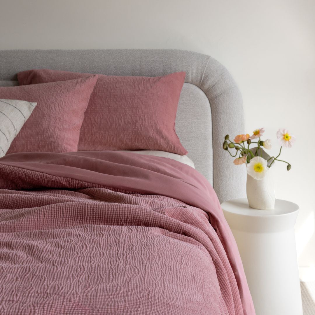 Bed with pink textured bedding and pillows, next to a small table with flowers.