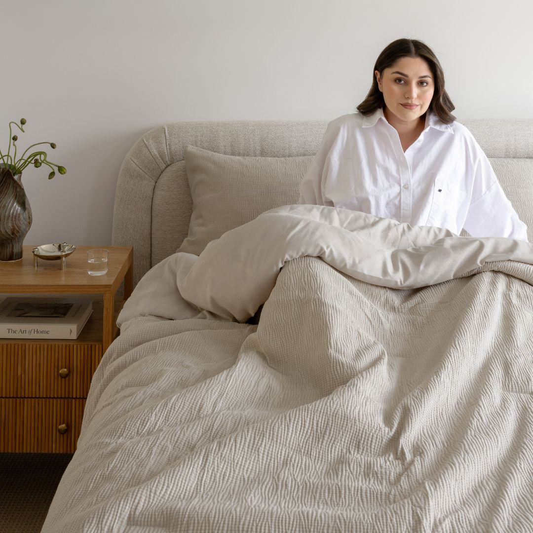 Woman in a white shirt sitting on a bed with beige bedding, next to a wooden nightstand.