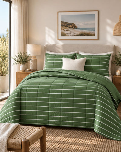 A green quilted comforter set with white stripes and matching pillow shams styled on a bed in a warm, sunlit bedroom. The scene includes a beige headboard, wooden nightstands, a lamp, a framed beach landscape print on the wall, and a woven bench in the foreground.
