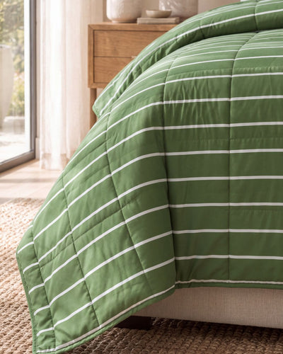A green quilted comforter with thin white horizontal stripes draped over a bed in a softly lit bedroom setting.