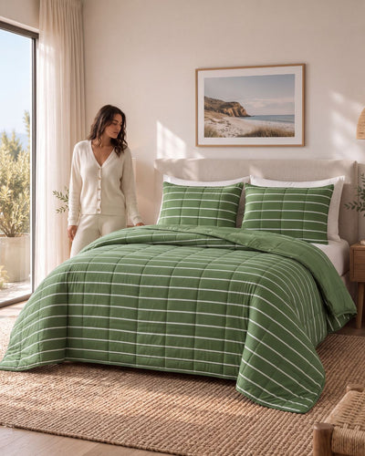 A woman in a cream cardigan and matching pants stands beside a bed styled with a green striped quilt and matching pillow shams in a bright, neutral-toned bedroom.