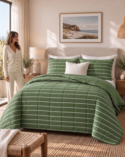 A woman in cream pajamas stands beside a bed styled with a green and white striped quilted comforter and matching pillow shams in a warm, sunlit bedroom.
