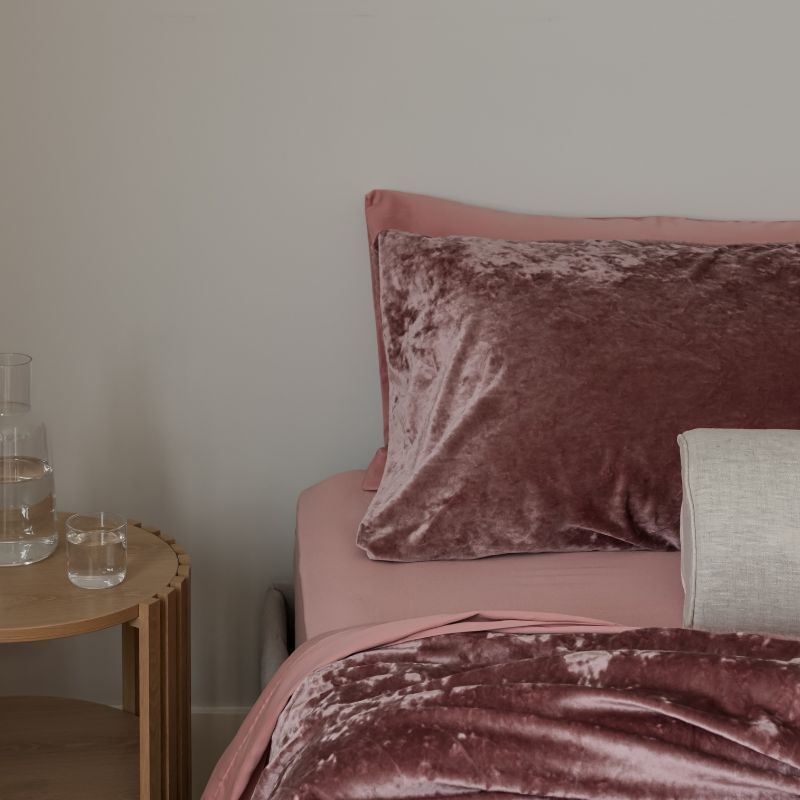 Bed with pink velvet bedding and pillows next to a wooden side table with a glass of water.
