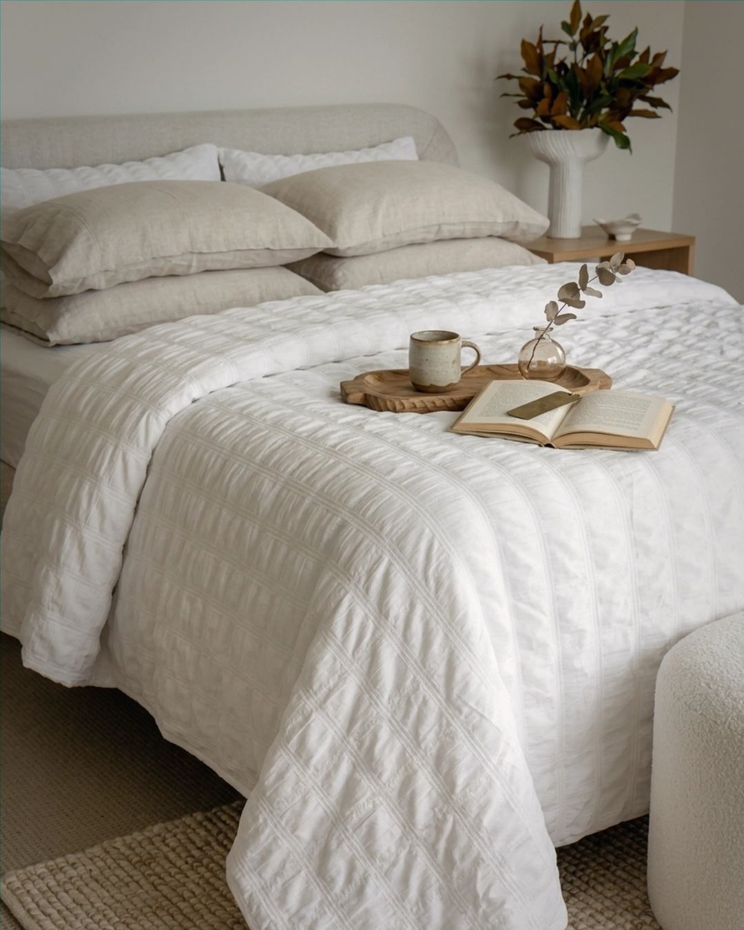 Neatly made bed with white bedding, a cup, book, and tray on a wooden nightstand.