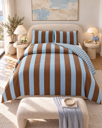 A bed dressed in a brown and light blue vertically striped bedding set, styled in a bright, modern bedroom with matching pillows, bedside tables, lamps, and a bench at the foot of the bed.