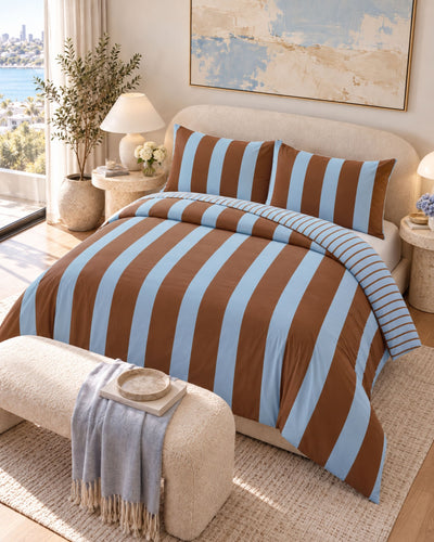 A bed dressed in a brown and light blue striped bedding set, shown in a styled bedroom with pillows, bedside tables, lamps, a bench, and a window view.