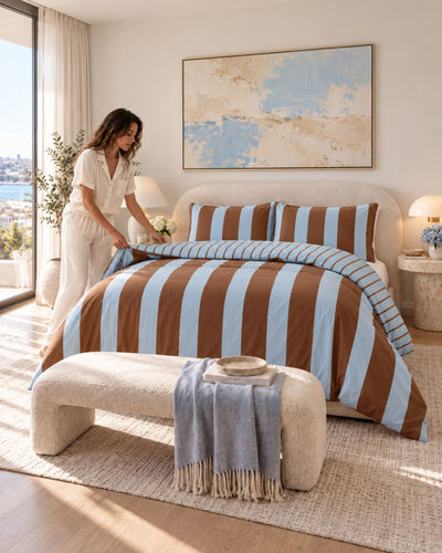 A woman in white loungewear adjusts a striped duvet cover on a bed in a bright, modern bedroom. The bedding features alternating light blue and brown vertical stripes, with matching striped pillows, a textured cream bench at the foot of the bed, and a blue throw blanket.