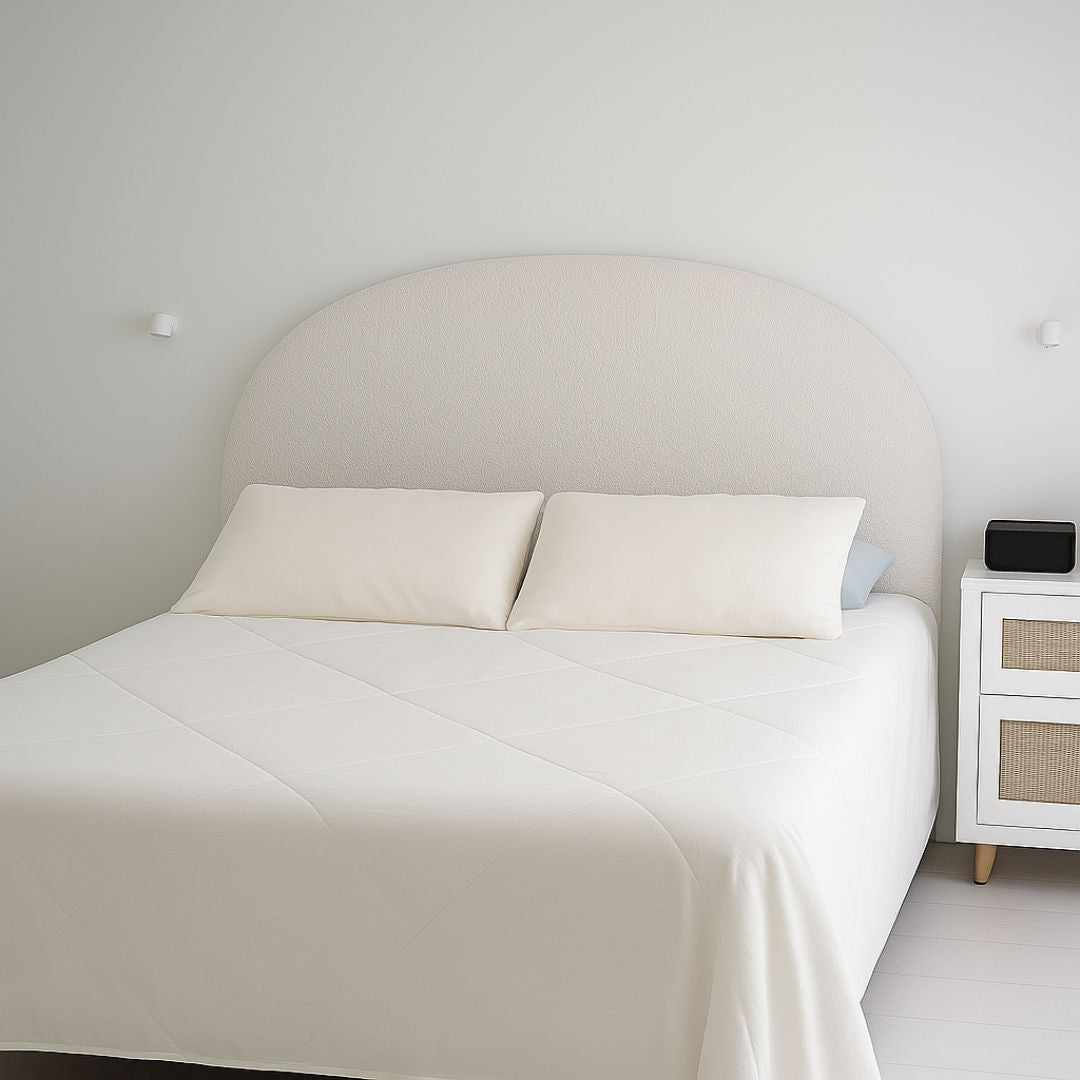 A minimalist bedroom features a neatly made bed with white bedding and two pillows, topped with the Sienna Living Summer Bamboo Cooling 2 in 1 Comforter Quilt. A lamp sits on a small nightstand, and sheer white curtains cover the window.