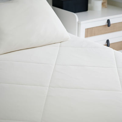 A minimalist bedroom features a neatly made bed with white bedding and two pillows, topped with the Sienna Living Summer Bamboo Cooling 2 in 1 Comforter Quilt. A lamp sits on a small nightstand, and sheer white curtains cover the window.