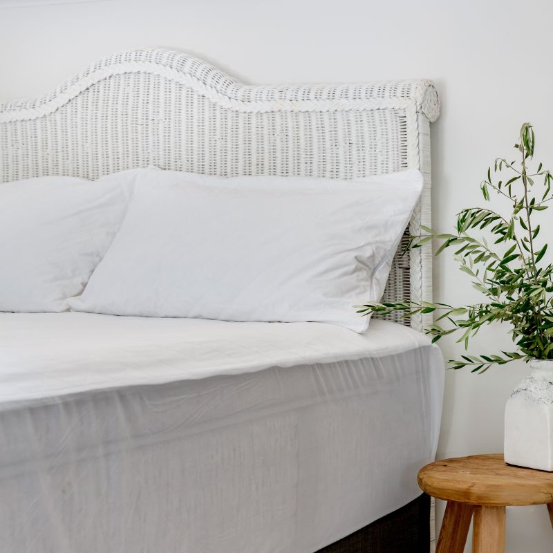 A neatly made bed with white sheets and pillows, a wicker headboard, and the Sienna Living Bamboo Mattress Protector. A small wooden table beside the bed holds a white vase filled with green leafy branches.