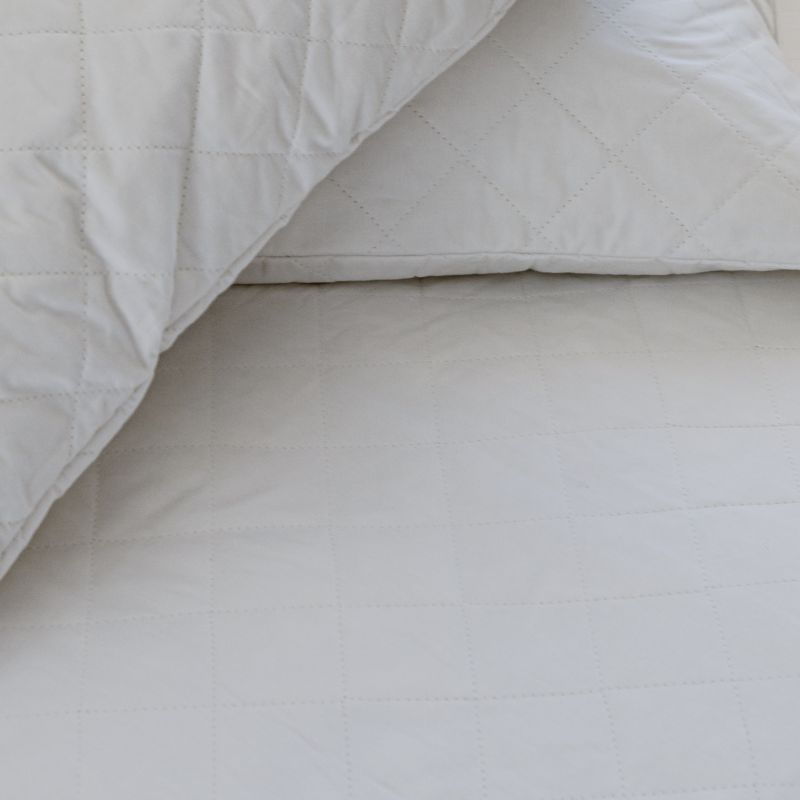 A neatly made bed with white sheets and pillows, a wicker headboard, and the Sienna Living Bamboo Mattress Protector. A small wooden table beside the bed holds a white vase filled with green leafy branches.