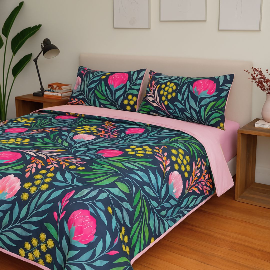 A bed styled with the Sienna Living BSM Claire Coverlet Set—featuring a dark floral pattern with pink flowers and green leaves—plus matching pillows, wooden nightstands, plants, and minimalist art on the white wall above.