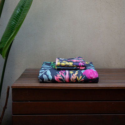 A bed styled with the Sienna Living BSM Claire Coverlet Set—featuring a dark floral pattern with pink flowers and green leaves—plus matching pillows, wooden nightstands, plants, and minimalist art on the white wall above.