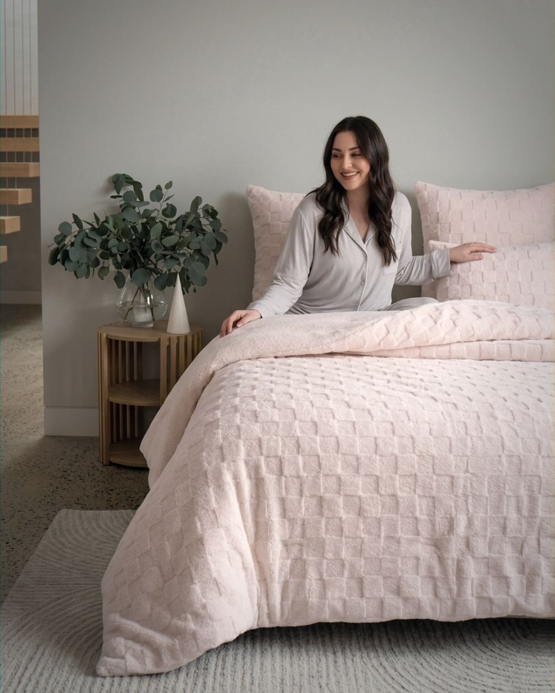 Lifestyle shot of woman in grey pajamas sitting on bed with soft pink checkered fleece comforter set
