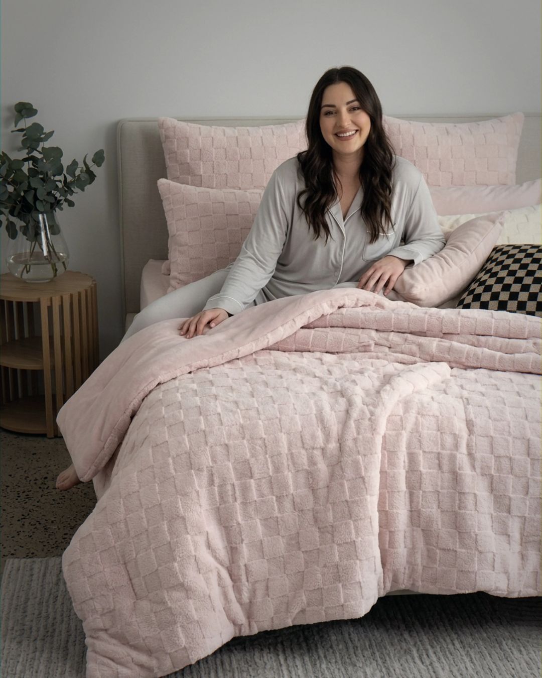 Lifestyle shot of woman sitting on bed with soft pink checkered fleece comforter set and matching pillows
