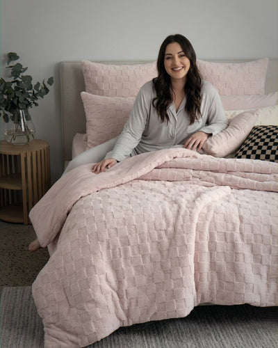 Lifestyle shot of woman sitting on bed with soft pink checkered fleece comforter set and matching pillows