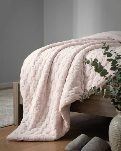 Soft pink checkered snuggle fleece comforter draped over bed foot, with eucalyptus plant and grey slippers nearby.