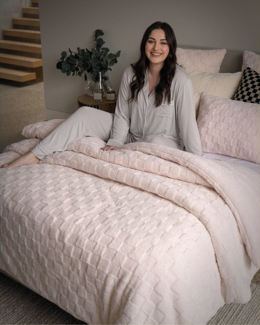 Lifestyle shot of woman in grey pajamas sitting on bed with soft pink checkered fleece comforter set