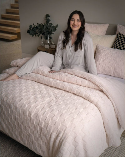 Lifestyle shot of woman in grey pajamas sitting on bed with soft pink checkered fleece comforter set